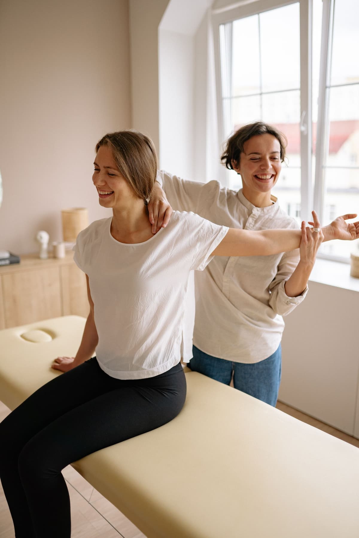 A physiotherapist guiding a smiling woman through a shoulder stretch in a bright, calm room