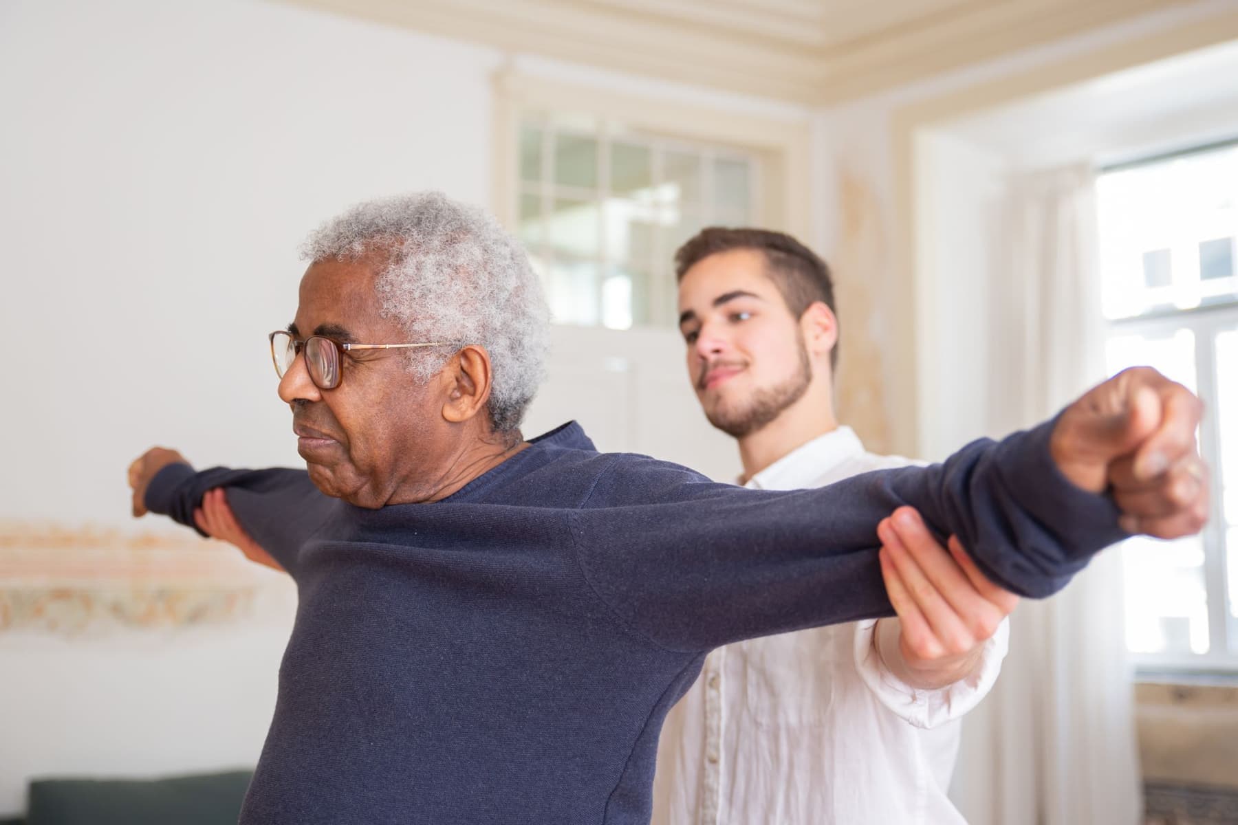 A physiotherapist supporting an older man's arm as he stretches in his living room